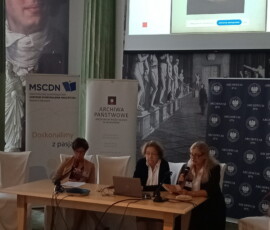Fotografia 3 kobiet siedzących za stołem prezydialnym. Nad stołem widoczny telebim z prezentacją. Jedna kobieta trzyma mikrofon.