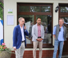 Fotografia mężczyzny stojącego z mikrofonem. W tle dwóch mężczyzn stojących przed witryną.