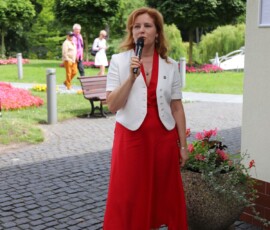 Fotografia kobiety w sukni z mikrofonem. W tle widoczne aleje parkowe w Międzyzdrojach.