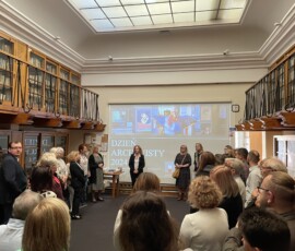 Fotografia tłumu osób zgromadzonych na sali w trakcie przemówienia kobiety. W tle widoczny telebim z prezentacją multimedialną.