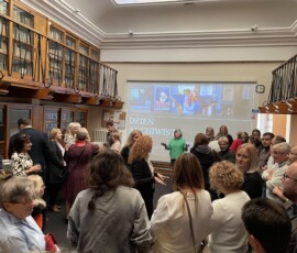 Grupa ludzi słucha kobiety. Z tyłu telebim z prezentacją.