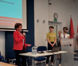 Fotografia 4 kobiet. Dwie stoją przy mównicy, jedna trzyma mikrofon. Widoczny stół z rzutnikiem i telebim na ścianie.