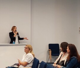Fotografia kobiety z mikrofonem mówiącej do siedzących słuchaczy.