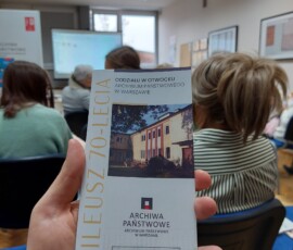 Zdjęcie dłoni z ulotką "Jubileusz 70-lecia" i kilku osób siedzących na sali, odwróconych plecami do fotografa.