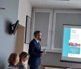Fotografia stojącego mężczyzny i dwóch kobiet w trakcie wystąpienia. Widoczny ekran z prezentacją multimedialną i grupa słuchaczy.