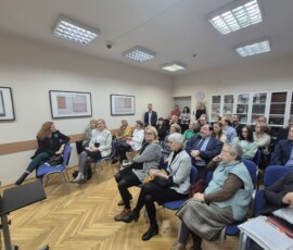 Fotografia grupy ludzi siedzących na krzesłach wewnątrz sali. Na ścianach wiszą obrazy w antyramach. Zdjęcie z lewego profilu.