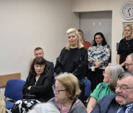 Fotografia kobiety w trakcie przemówienia. Dookoła siedzą ludzie na krzesłach wewnątrz sali. Zdjęcie z dali.