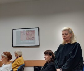 Fotografia kobiety w trakcie przemówienia. Dookoła siedzą ludzie na krzesłach wewnątrz sali.