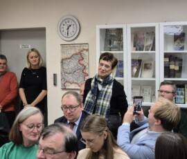 Fotografia kobiety w trakcie przemówienia. Dookoła siedzą ludzie na krzesłach wewnątrz sali. Zdjęcie z dali.