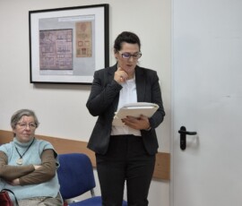 Fotografia kobiety w marynarce z mikrofonem. Dookoła siedzący słuchacze wewnątrz sali.