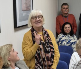 Fotografia kobiety z mikrofonem z żółtej bluzce i słuchaczy wewnątrz sali.