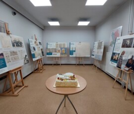 Fotografia stolika z tortem, stojącego na środku sali. Dookoła stolika plansze wystawiennicze na sztalugach, ułożone w prostokąt.
