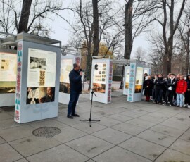 Łukasz Nowacki - specjalista do spraw historii miasta w Ośrodku Kultury Gminy Grodzisk Mazowiecki, fot. M. Goroch Fotografia kolorowa mężczyzny w okularach stojącego przed mikrofonem na zewnątrz. Widoczne kolorowe plansze wystawowe i drzewa.