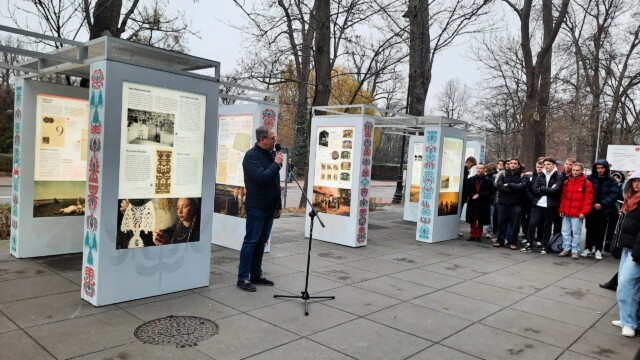 Łukasz Nowacki - specjalista do spraw historii miasta w Ośrodku Kultury Gminy Grodzisk Mazowiecki, fot. M. Goroch Fotografia kolorowa mężczyzny w okularach stojącego przed mikrofonem na zewnątrz. Widoczne kolorowe plansze wystawowe i drzewa.