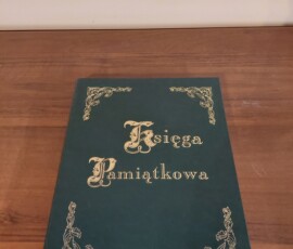 Fotografia kolorowa księgi w zielonej okładce, leżącej na drewnianym stole.