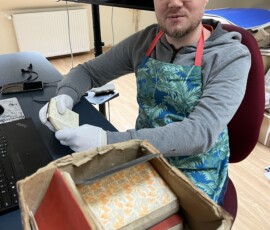 Adrian Kossowski w trakcie porządkowania archiwalnej Spuścizny po prof. Istvánie Lagzim, fot. A. Szczęsnowicz-Panas Fotografia kolorowa mężczyzny w okularach, siedzącego przy biurku. Widoczne stare dokumenty w pudle.