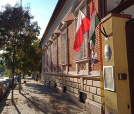 Kolorowa fotografia budynku z flagą biało-czerwoną i flagą biało-czerwona-zieloną. Widoczny chodnik i samochody.