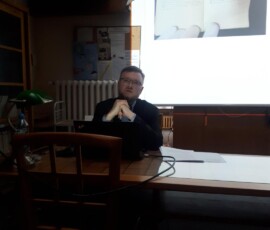 Fotografia ciemna, kolorowa z mężczyzną patrzącym w górę w pozycji siedzącej. W tle płachta z wyświetlaną prezentacją.