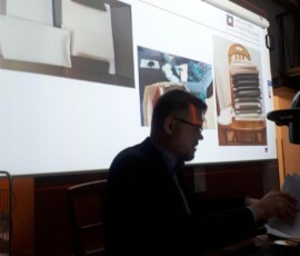 Fotografia ciemna, kolorowa z mężczyzną patrzącym w górę w pozycji siedzącej. z prawego profilu W tle płachta z wyświetlaną prezentacją.