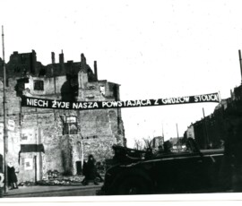Transparent na paradę z okazji rocznicy wyzwolenia Warszawy, styczeń 1946 r. Na czarno-białej fotografii widać ruiny budowli, transparent z napisem „Niech żyje nasza powstająca z gruzów stolica!”, samochód oraz przechodzących ulicą ludzi.