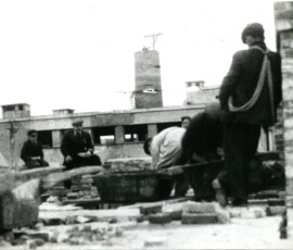 Odbudowa III kolonii Warszawskiej Spółdzielni Mieszkaniowej przy ul. Suzina, październik 1945 r. Na czarno-białej fotografii widać sześciu mężczyzn odpoczywających wśród cegieł, w tle budowana wieża.