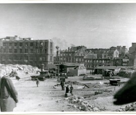 Rozbiórki ruin na Mariensztacie, kwiecień 1948 r. Na czarno-białej fotografii widać zamyślonego mężczyznę, w tle grupa mężczyzn, sprzęt budowlany i ruiny budynków.
