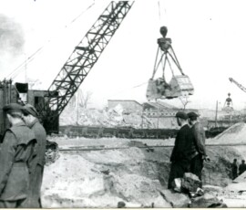 Roboty przy wykopie pod tunel pod Krakowskim Przedmieściem, kwiecień 1948 r. Na czarno-białej fotografii widać grupę kilku mężczyzn, w tle sprzęt budowlany, budynek z kominem i ruiny innych budynków.