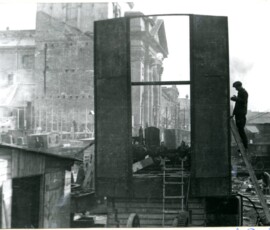 Prace na Pl. Zamkowym i Krakowskim Przedmieściu, kwiecień 1948 r. Na czarno-białej fotografii widać robotnika na drabinie przy metalowej konstrukcji, obok drewniany schowek, a w tle sprzęt budowlany i budynek.