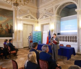 Fotografia kolorowa. Kobieta stoi przed mikrofonem z bordową teczką. W tle widać siedzących słuchaczy.