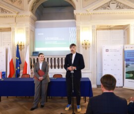 Fotografia kolorowa. Mężczyzna z mikrofonem. Kobieta stoi z bordową teczką.