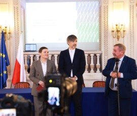 Fotografia kolorowa. Mężczyzna stoi przy mikrofonie. Obok stoją mężczyzna i kobieta.