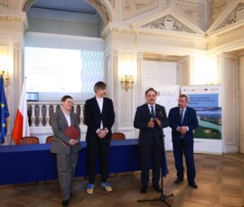 Fotografia kolorowa. Kobieta i trzech mężczyzn stoi z dokumentem w bordowej obwolucie. Za nimi stół z niebieskim obrusem. W tle flaga Polski i Unii Europejskiej.