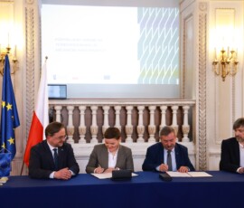Fotografia kolorowa. Kobieta i trzech mężczyzn podpisuje dokument w bordowej obwolucie na stole z niebieskim obrusem. W tle flaga Polski i Unii Europejskiej.