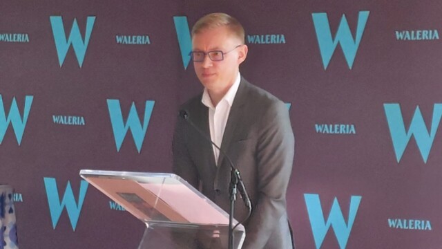 Marcin Dawidowicz, Mazowiecki Konserwator Zabytków w czasie przemowy na uroczystym otwarciu, fot. M. Aleksiejuk Fotografia kolorowa mężczyzny w okularach, stojącej na mównicy z mikrofonem. W tle bordowa plansza z błękitnymi literami.
