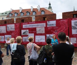 Otwarcie wystawy, na murach miejskich wiszą czerwone plansze wystawy z białymi rysunkami niezrealizowanych projektów warszawskich budynków