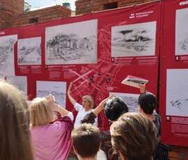 Otwarcie wystawy, na murach miejskich wiszą czerwone plansze wystawy z białymi rysunkami niezrealizowanych projektów warszawskich budynków