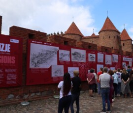 Otwarcie wystawy, na murach miejskich wiszą czerwone plansze wystawy z białymi rysunkami niezrealizowanych projektów warszawskich budynków. Przed murami grupa ludzi