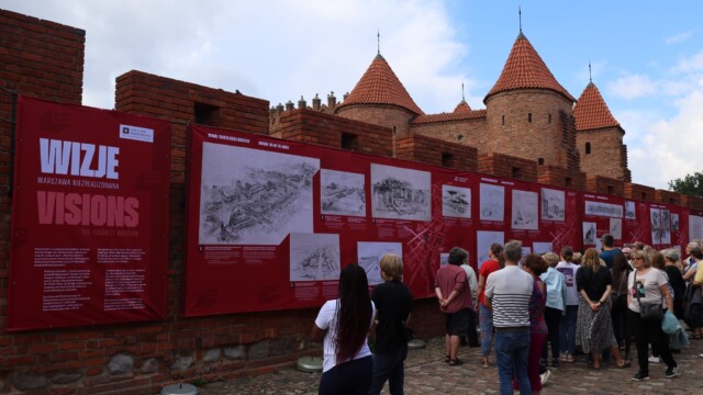 Otwarcie wystawy, na murach miejskich wiszą czerwone plansze wystawy z białymi rysunkami niezrealizowanych projektów warszawskich budynków. Przed murami grupa ludzi