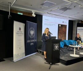 Zastępca dyrektora Archiwum Państwowego w Warszawie ds. zasobu i działalności archiwalnej Anna Wajs na IX Ogólnopolskiej Konferencji Archiwistów i Historyków z cyklu „Educare necesse est…”. Na kolorowej fotografii widać kobietę przy mikrofonie przed pulpitem, w tle banery z logotypami Archiwum Państwowego w Warszawie i Archiwum Instytutu Pamięci Narodowej oraz prezentacja multimedialna, jak również jeszcze jedna siedząca kobieta.