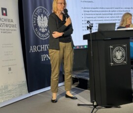 Magdalena Masłowska, kierownik Oddziału III Udostępniania Zasobu Archiwalnego w Archiwum Państwowym w Warszawie, na IX Ogólnopolskiej Konferencji Archiwistów i Historyków z cyklu „Educare necesse est…”. Na kolorowej fotografii widać kobietę z mikrofonem obok pulpitu, w tle banery z logotypami Archiwum Państwowego w Warszawie i Archiwum Instytutu Pamięci Narodowej oraz prezentacja multimedialna.