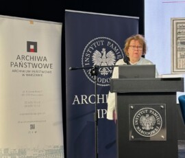Starszy kustosz Marta Jaszczyńska z Archiwum Państwowego w Warszawie na IX Ogólnopolskiej Konferencji Archiwistów i Historyków z cyklu „Educare necesse est…”. Na kolorowej fotografii widać kobietę przy mikrofonie przed pulpitem, w tle banery z logotypami Archiwum Państwowego w Warszawie i Archiwum Instytutu Pamięci Narodowej.