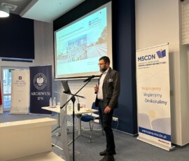 Kustosz Maciej Aleksiejuk z Archiwum Państwowego w Warszawie na IX Ogólnopolskiej Konferencji Archiwistów i Historyków z cyklu „Educare necesse est…”. Na kolorowej fotografii widać mężczyznę przy mikrofonie przed pulpitem, w tle prezentacja i banery z logotypami Archiwum Państwowego w Warszawie, Archiwum Instytutu Pamięci Narodowej i Mazowieckiego Samorządowego Centrum Doskonalenia Nauczycieli.