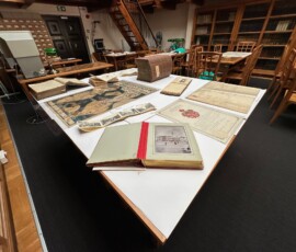 Materiały archiwalne i biblioteczne wykorzystane przy okazji lekcji archiwalnej w Archiwum Państwowym w Warszawie Na kolorowej fotografii widać rozłożone na stole księgi, albumy i dokumenty, w tle stoły, krzesła i szafy