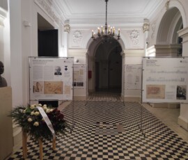 Wystawa „Warszawa silniejsza od wojny” w Gmachu Głównym Politechniki Warszawskiej. Na kolorowej fotografii widać plansze wystawiennicze, popiersie mężczyzny, wieniec i eleganckie wnętrze.