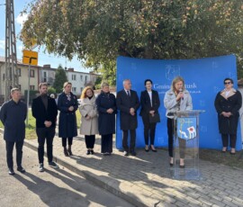 Przemawia Danuta Łada, Zastępca Kierownika Wydziału Inwestycji Urzędu Miejskiego w Pułtusku. Barwna fotografia przedstawiająca dziewięć osób na tle niebieskiej ścianki promocyjnej z napisem Gmina Pułtusk oraz drzew i budynków