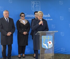Przemawia Pani Burmistrz Pułtuska Beata Jóźwiak Na zdjęciu widać kobietę z mikrofonem przy pulpicie, w tle dwie kobiety i mężczyzna oraz baner z napisem Gmina Pułtusk i herbem miasta.