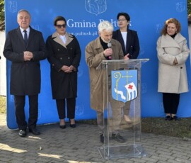 Przemawia Marek Gierlach, współautor projektu kompozycji ceramicznych płaskorzeźb Barwna fotografia przedstawiająca pięć osób na tle niebieskiej ścianki promocyjnej z napisem Gmina Pułtusk