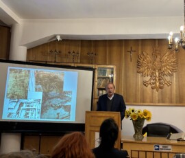 Wykład „O niegdysiejszej archeologii w Pułtusku” archeologa Zbigniewa Polaka Na zdjęciu widać mężczyznę przy pulpicie obok prezentacji, w tle drewniany orzeł i krzyż.