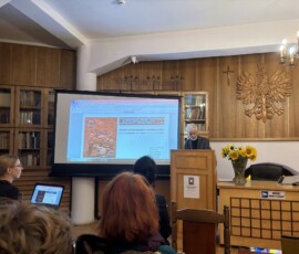 Wykład „O potrzebie miejsca zadumy” współautora projektu kompozycji, archeologa i historyka sztuki Marka Gierlacha. Na zdjęciu widać mężczyznę przy pulpicie obok prezentacji, w tle drewniany orzeł i krzyż.