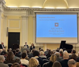 Przemawia Pani Magdalena Wiercińska, dyrektor Archiwum Państwowego w Warszawie. Na kolorowej fotografii widać przemawiającą do grupy ludzi kobietę, w tle prezentacja, fortepian i sprzęt komputerowy.